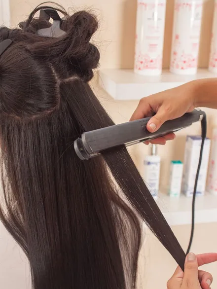 Estilista planchando un mechón de cabello oscuro mechón por mechón, con el resto del pelo recogido y productos profesionales en el estante del fondo